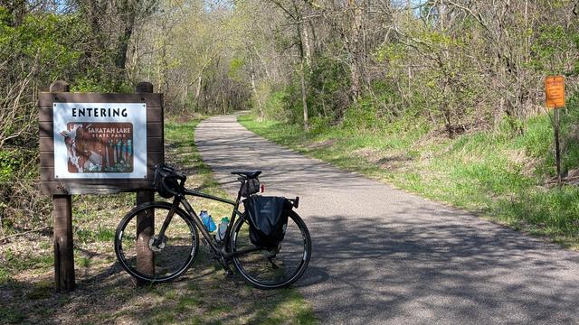 Dark brown Diverge bicycle leaning against the wooden sign for "Entering Sakata Lake State Park" that has a Pileated Woodpecker and wildflowers painted on it. To the right the asphalt trail disappears after a curve to the right with newly leafing trees and shrubs on either side,