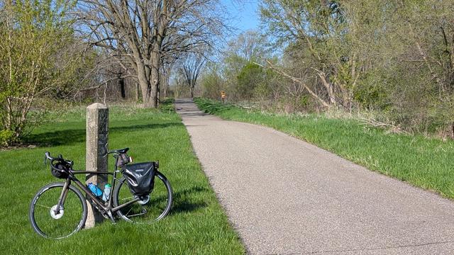 Dark brown Diverge bicycle leaning on the concrete obelisk Chicago Great Western Milepost "M43" on the green lawn to the left of the Sakatah Singing Hills State Trail with newly leafing trees and shrubs on either side.