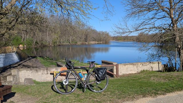 Dark brown Diverge bicycle leaning on a bike rack in front of the dam that forms Lake Louise from the Upper Iowa River. The lake is surround by trees leafing out and the water reflects the clear blue sky.