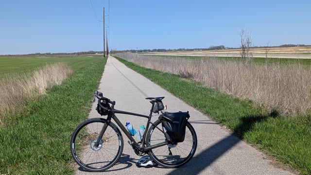 Dark brown Diverge bicycle parked on the asphalt Shooting Star State Trail which goes straight to the vanishing point. On the left are power utility poles and farm fields and on the right is MN Highway 56 and the long grass in the ditch in between. The water tower for Rose Creek can barely be seen in the distance.