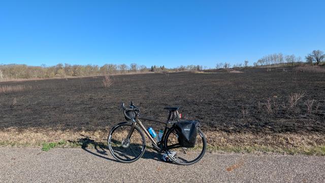 Dark brown Diverge bicycle parked on the Shooting Star State Trail in Lake Louise State Park in front of a prairie that has recently undergone a prescribed burn and looks mostly black with little bits of green foliage emerging under blue skies. There is a line of trees in the distance.