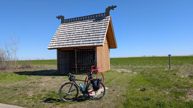 Dark brown Diverge bicycle leaning on a red bike shaped bicycle rack in front of the Norwegian Hill rain shelter on the Shooting Star State Trail. The shelter has Viking dragon heads on the top and steep wood shake roof. It is surrounded by green foliage under blue skies.