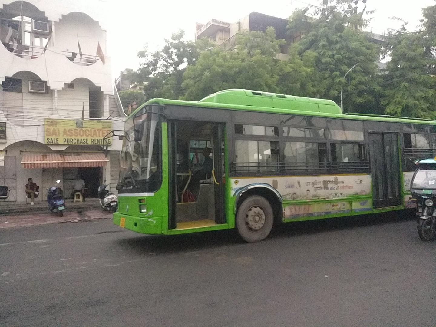 Ashok Leyland Non Airconditioned Ultra Low Floor City bus with some advertisment on its side gliding down the street with its front exit door still open with an e rickshaw right behind it