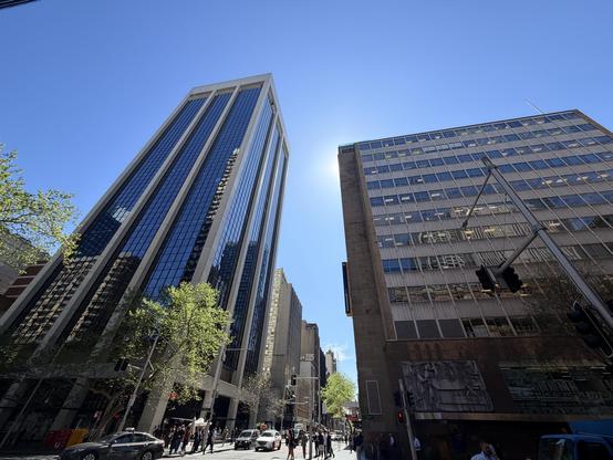 An ultra-wide angle photo of the street on a clear blue sky day (after rain), starting just below the horizon to include the street and cars turning in front a building as a tiny proportion of the photo — but cropping pedestrians within a few metres.  The sun is subtly evident just behind the corner of a building, such that walking one second in either direction would reveal it.  The top of the photo’s ultra-wide angle makes it all the way up two office buildings, one of which is a 20-25 floor …