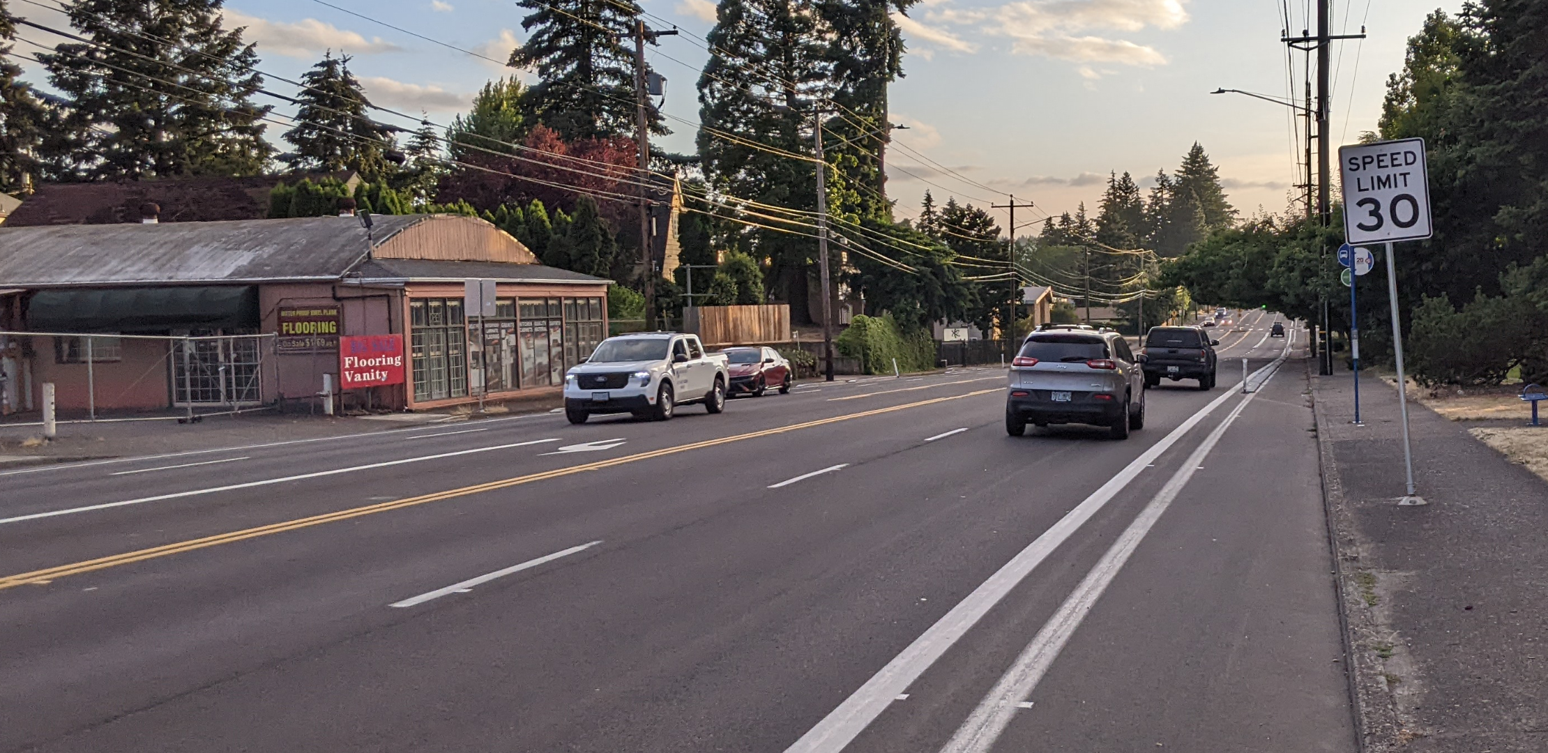 Five lane stroad with buffered and sometimes curb-protected bike lanes, curb-tight sidewalks, sun setting other conifer trees and Mt Tabor.