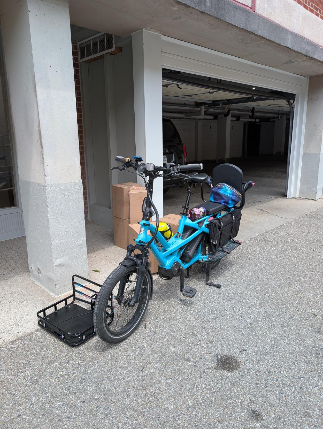 Blue long-tail cargo bike in front of a building garage, with a front rack on the ground and a stack of boxes behind