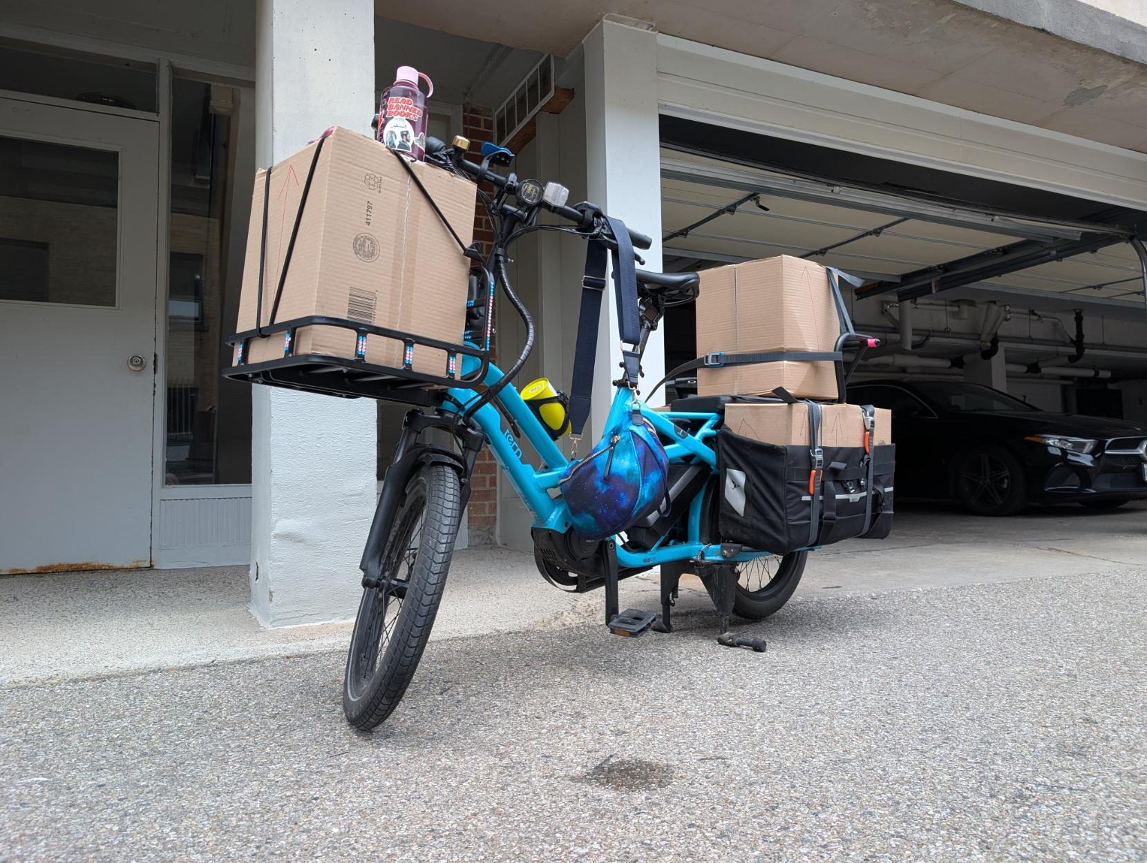 A blue long-tail cargo bike in front of a building garage, with a front rack loaded with a box, a box in the back seat, and boxes in the panniers
