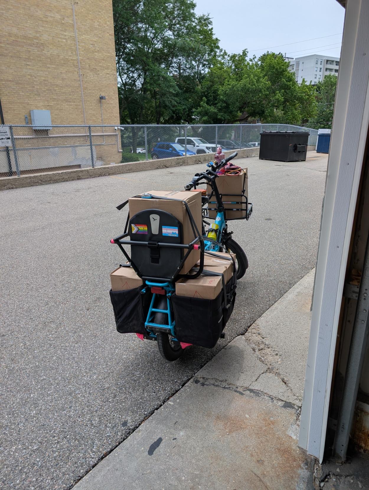 A shot from behind of a blue long-tail cargo bike in front of a building garage, with a front rack loaded with a box, a box in the back seat, and boxes in the panniers. Pride stickers are visible on the back of the back seat, progress pride and trans pride