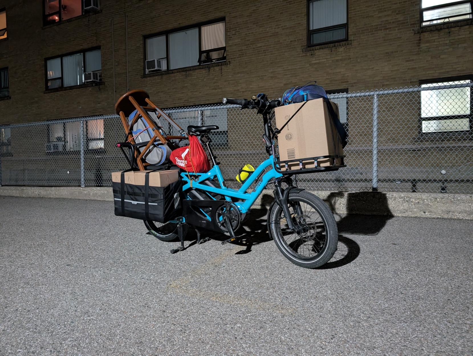Night time, side view, a fully loaded blue long-tail cargo bike with a front rack, with a building behind. There are boxes in the panniers and a a box on the front rack, and a stool perched in the back with a trumpet case inside