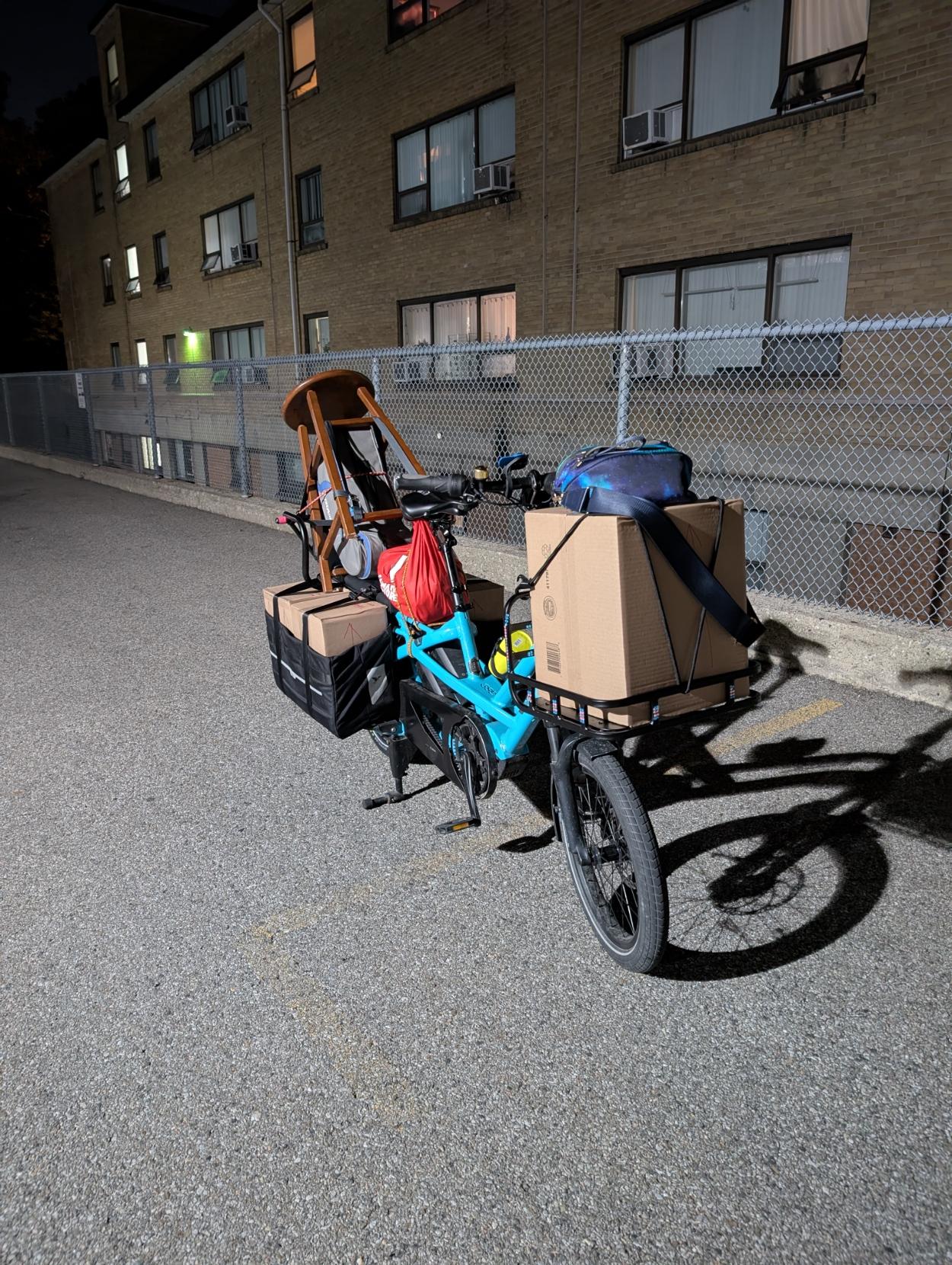 Night time, 3/4 view, a fully loaded blue long-tail cargo bike with a front rack, with a building behind. There are boxes in the panniers and a a box on the front rack, and a stool perched in the back with a trumpet case inside
