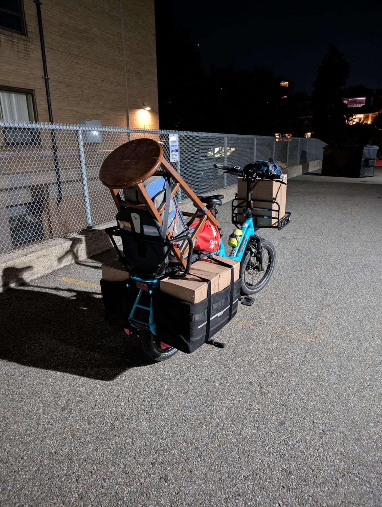 Night time, rear view, a fully loaded blue long-tail cargo bike with a front rack, with a building behind. There are boxes in the panniers and a a box on the front rack, and a stool perched in the back with a trumpet case inside