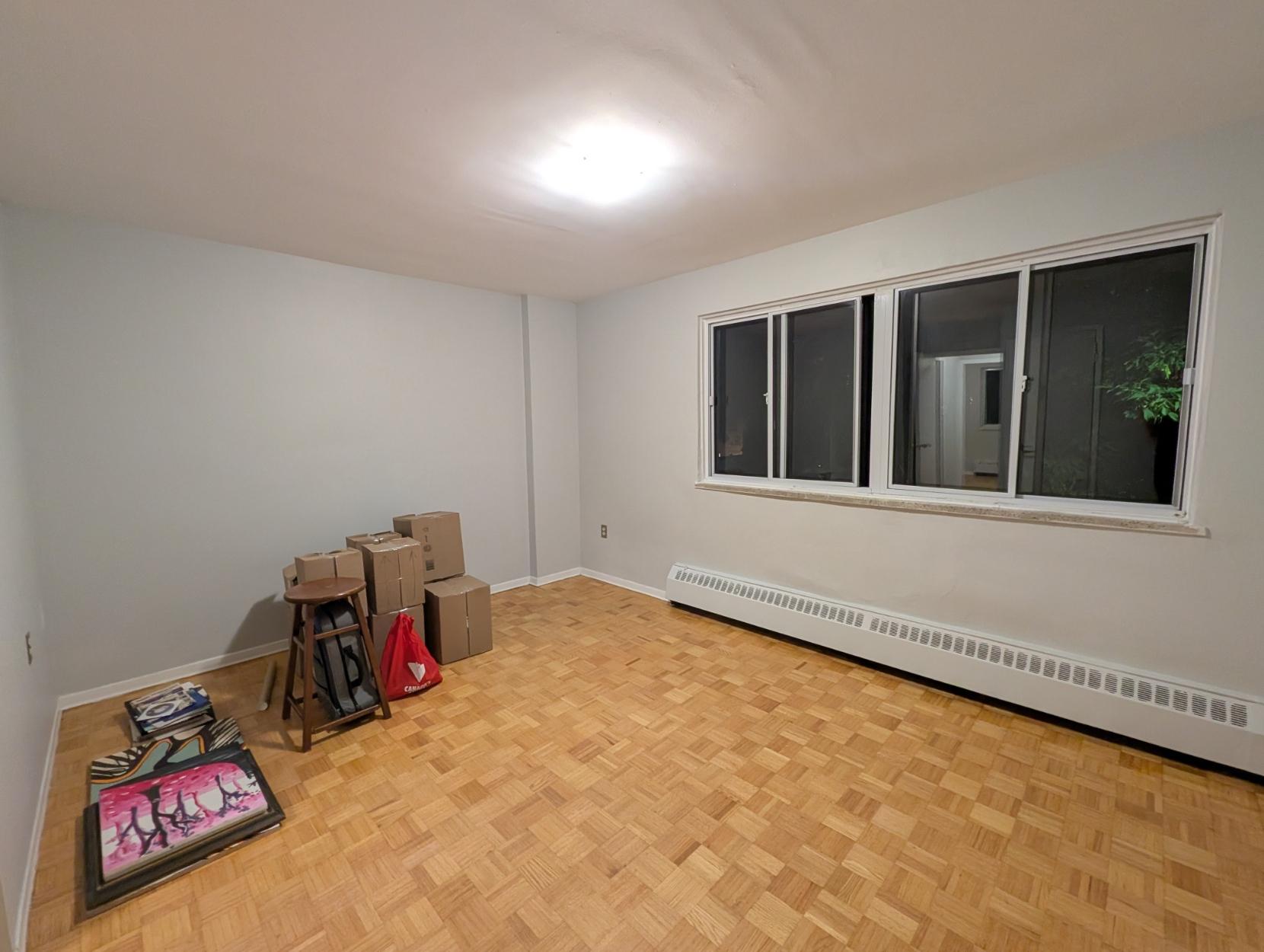 A room viewed from the corner, a wide window and baseboard heater on one wall and an assortment of boxes along the other