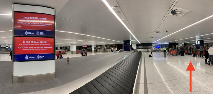 A tight panorama of the baggage carousel, showing Derryn Hinch in the distance, waiting to the right at the next carousel and talking to people, while waiting for baggage.  Close up, on the left of the photo can be seen the sexual offences advertisement that explained the laws he had contributed to.