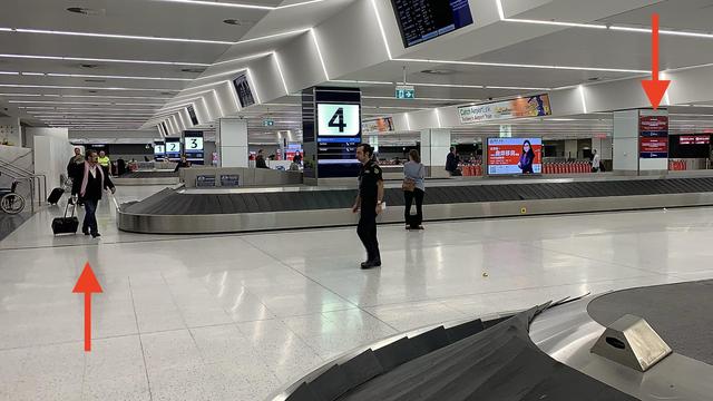 A long and wide shot of the baggage carousel, showing Derryn Hinch at the next carousel walking by with his carry-on luggage.  At the other end of the photo can be seen the sexual offences advertisement that explained the laws he had contributed to.