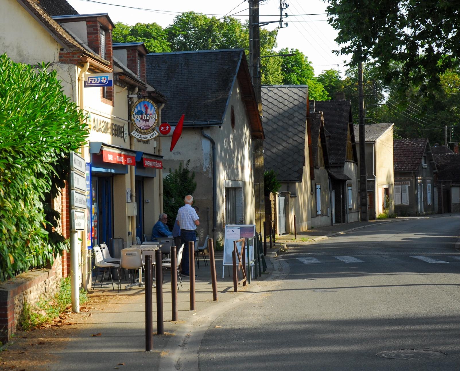 A small Bar Tabac at the main intersection in an old house. A few tables and chairs outside, shaded by a very old tree. Two locals having a chat.