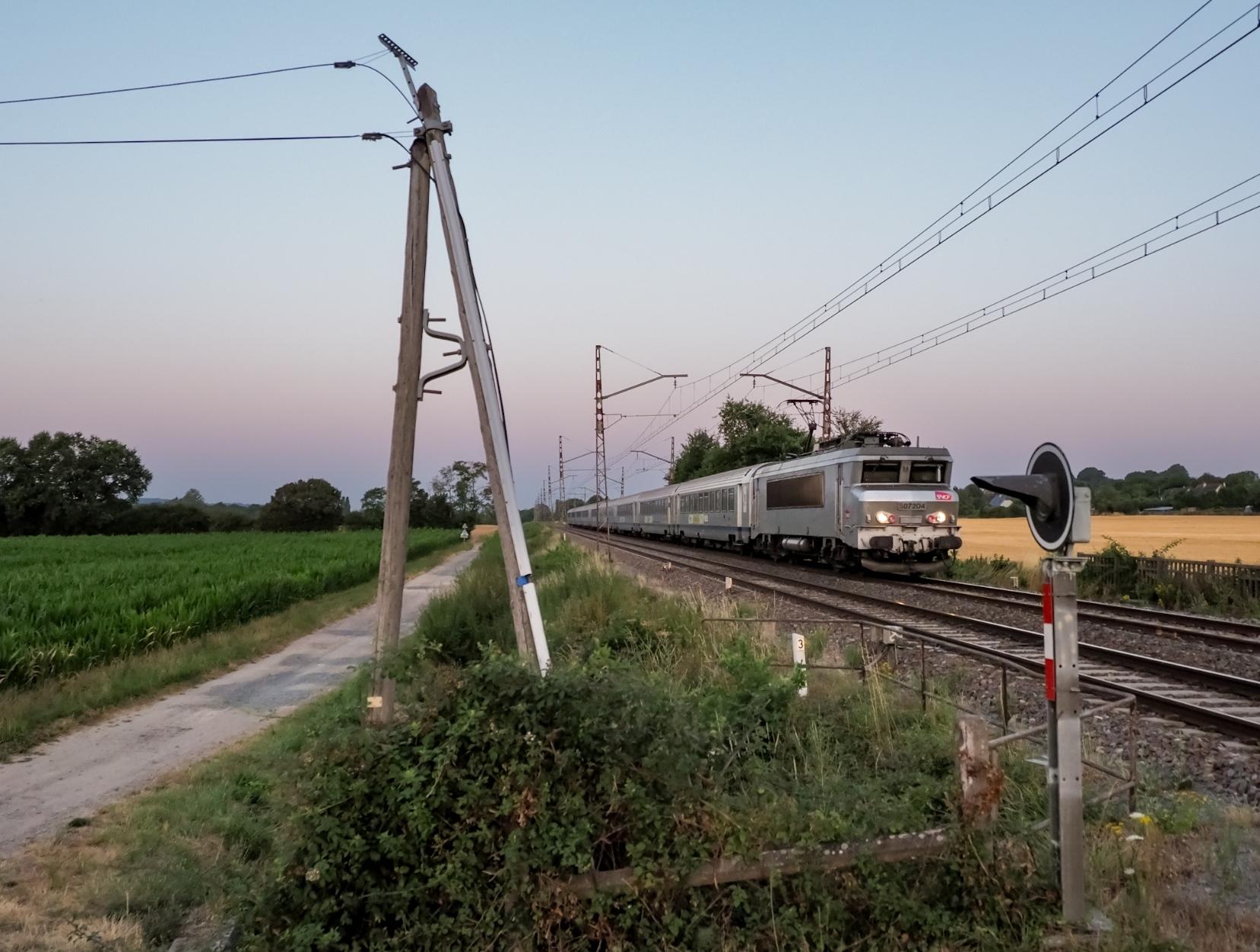 In grey-ish morning light just before sunrise, a silver fantôme liveried Nez Cassé hauls a long rake of Corail carriages past an old telephone or electricity pole.