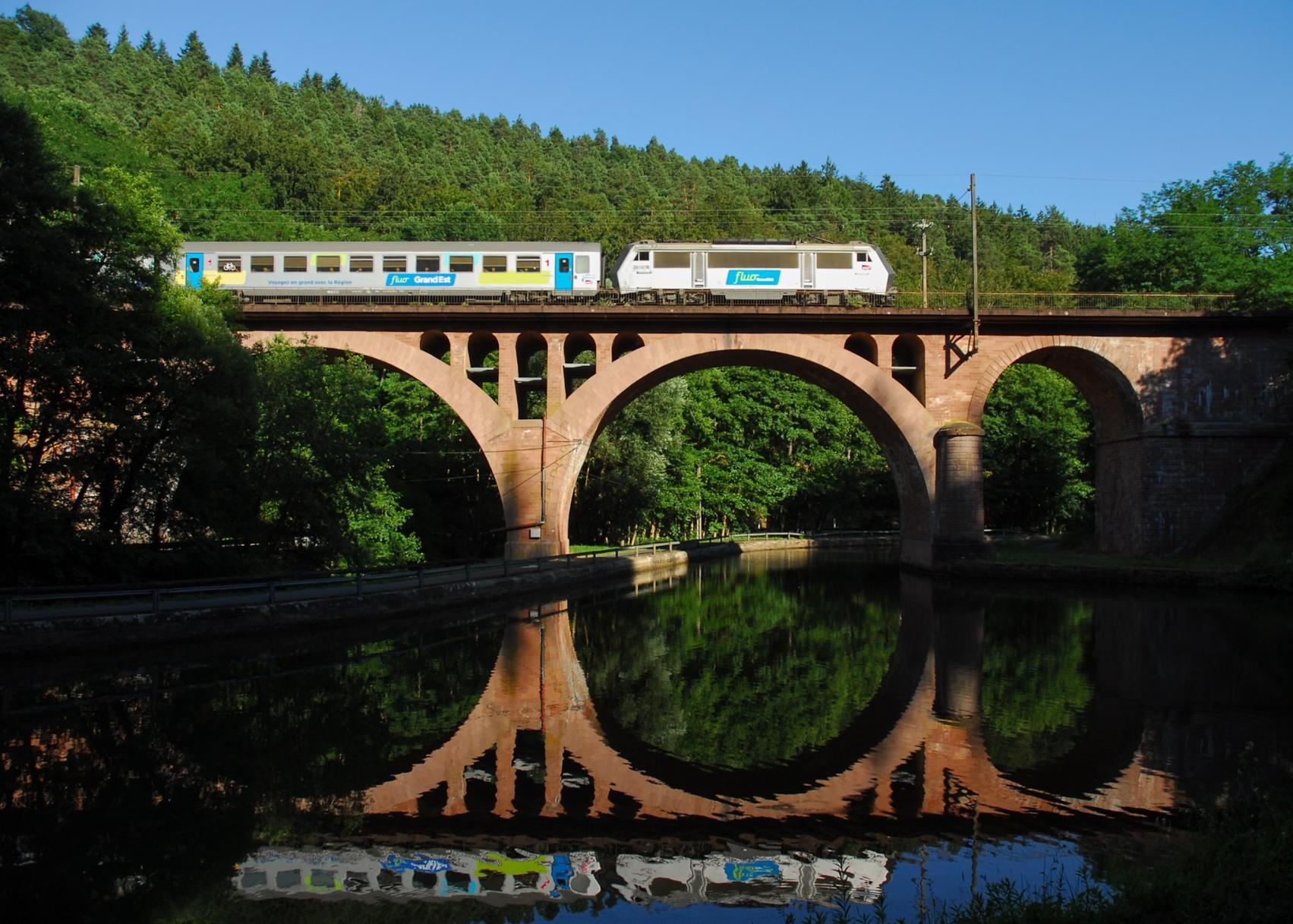 A stone arch viaduct spans a slightly rippled Canal in the middle of a forest in a valley. A silver "Fluo" liveried Sybic with a rake of Corail carriages, of which only the first is visible, crosses the bridge.