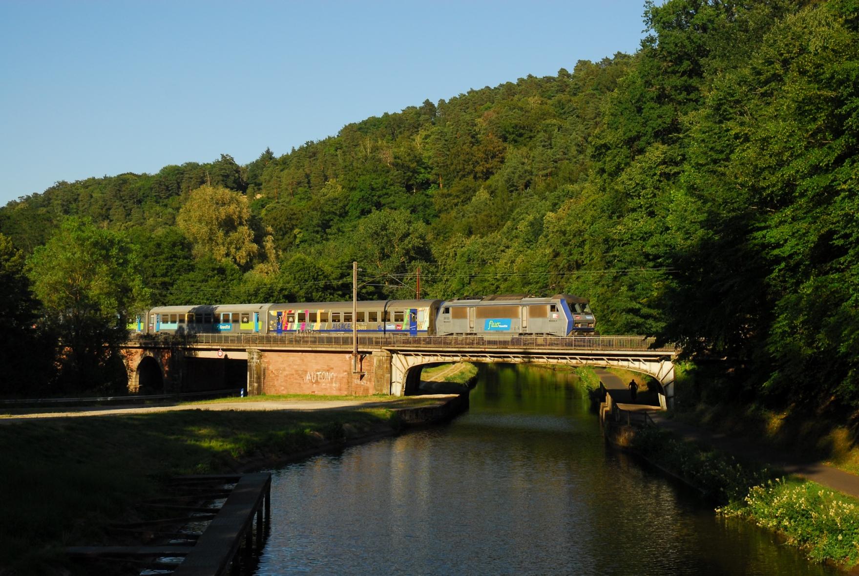 A low bridge spanning the canal and a nearby road in a valley. All surrounded by forest. A Sybic with a Corail train passes the bridge in warm evening light.