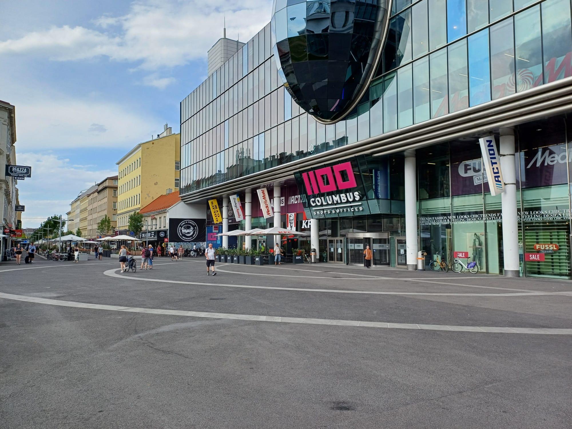 Ein großer, schwarz asphaltierter Platz bei sonnigem Wetter. Rechts das Einkaufszentrum mit einer Glasfassade. Der Platz ist so gut wie verwaist, da er sich in der Hitze stark aufheizt.