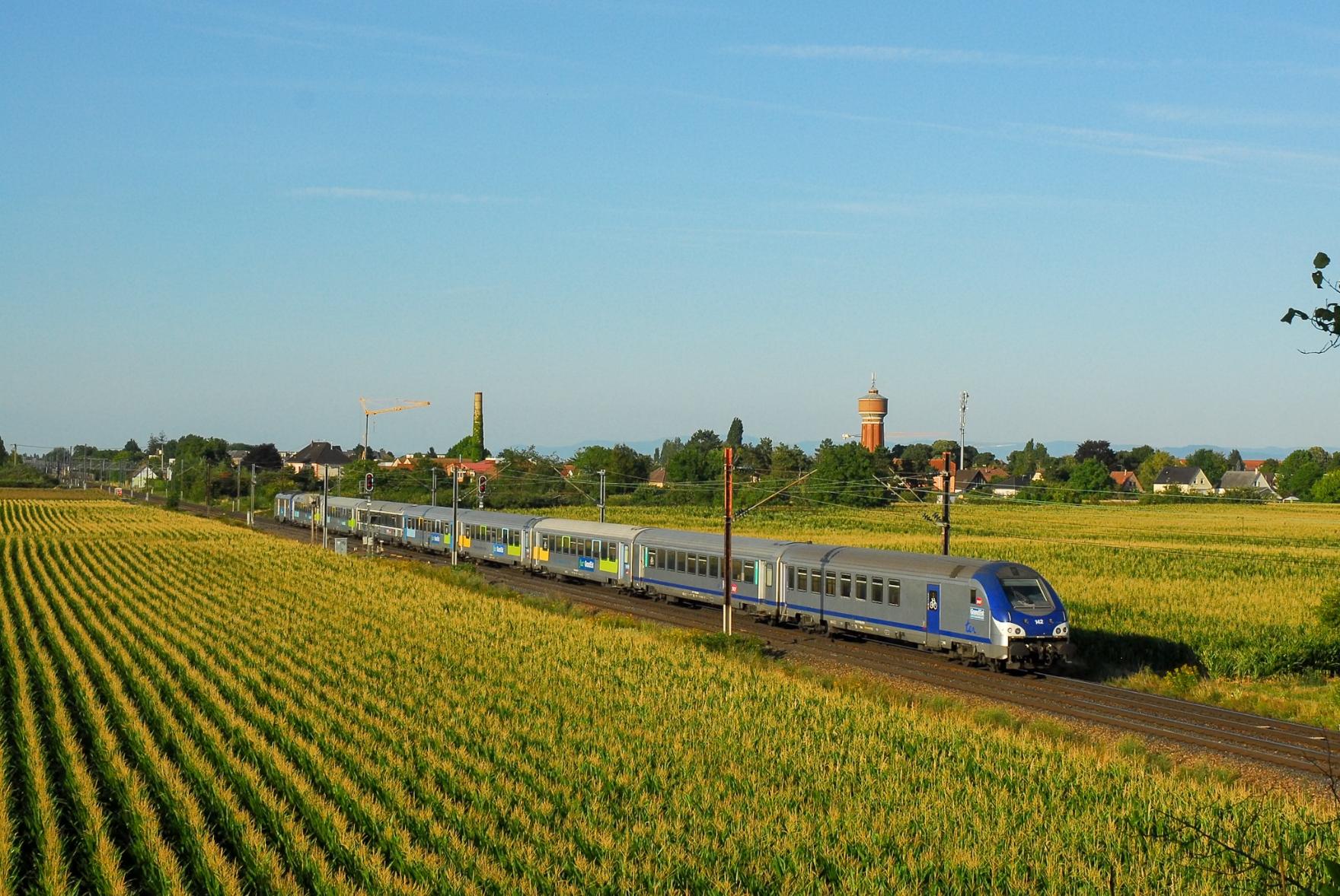 A Corail train with mixed liveries on a straight strech of railway main lines across cornfields. The Sybic loco is at the very end (well, actually: the front) of the train, the driving trailer in classic generic TER livery points towards the camera. Behind the train in the distance is a chimney and an old water tower of a nearby village. Warm evening light, although a bit dimmed by high clouds.