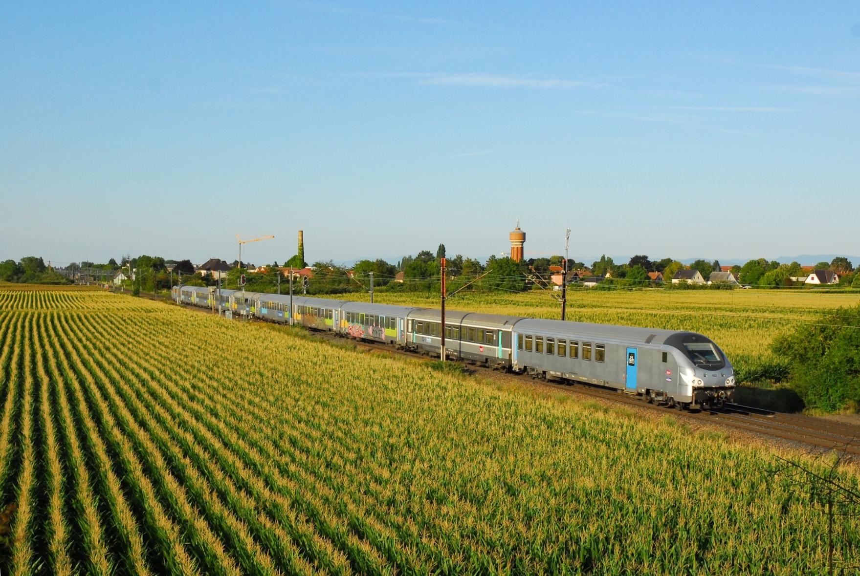 A TER 200 train in the landscape: Driving trailer in livrée fantôme, a carriage in the old "Corail+" colours and then 7 carriages with "Fluo" stickers, some of them heavily tagged. A Sybic locomotive at the rear. The cornfields reflect the warm summer evening light. A village with a brick water tower and an old chimney as landmarks in the background.