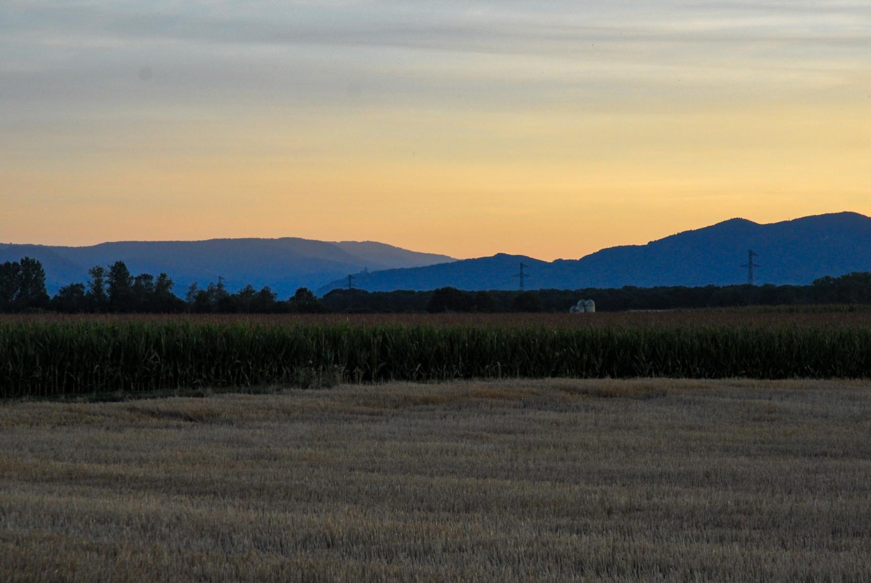 View across the flat landscape towards the Voges mountains after sunset. The hills appear layered in the distance. A pastel orange colour in the sky.