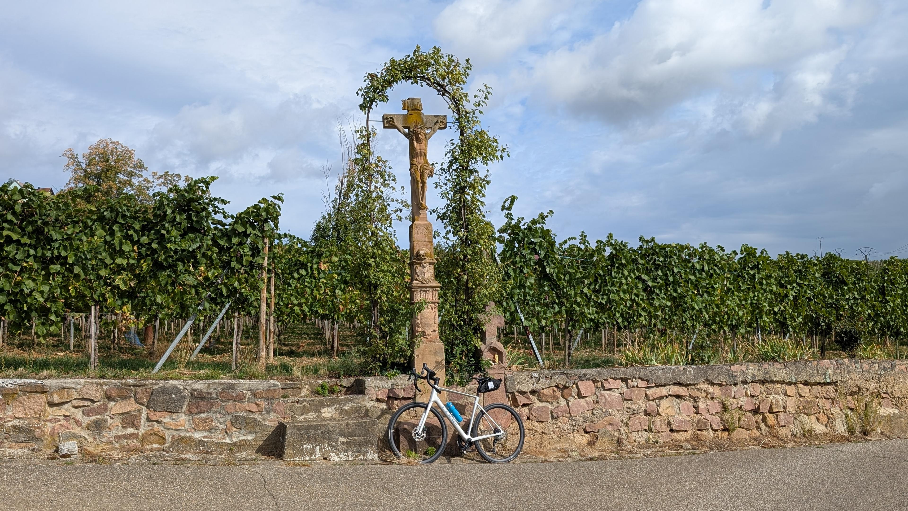 A bike parked in front of a stone crucifix in front of a vineyard.