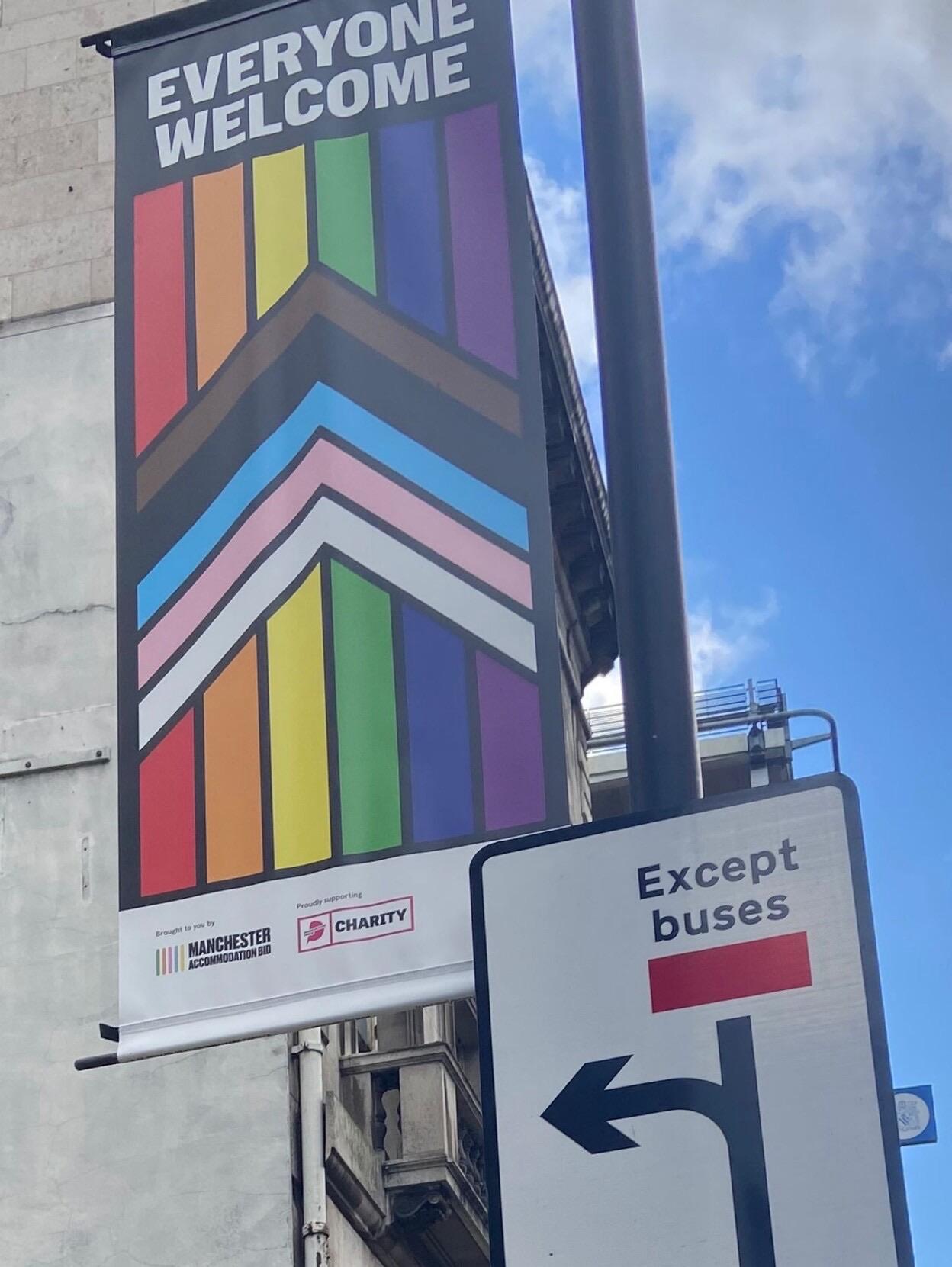 A picture of a BIPOC LGBTQ flag hanging on a lamp post with the words “EVERYONE WELCOME”. Below the flag is a sign for a bus lane, which is cropped so the only visible words are “Except buses” 