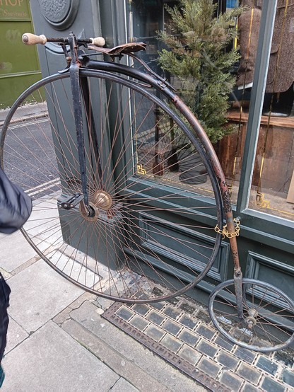 Penny farthing bike chained to a store. At the edge of the picture you can see there is someone just of camera.