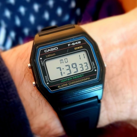 An on wrist black (plastic) Casio F-84W digital watch with a couple of prominent scratches on the face. The time shown is 7:39:33 on Monday the 11th.