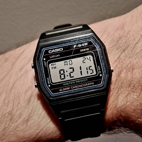 A black (plastic) Casio F-84W digital watch on wrist with many scratches on the face. The time shown is 8:21:15 on Monday the 24th.
