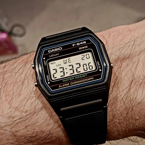 A black (plastic) Casio F-84W digital watch on wrist with without any scratches on the face. The time shown is 23:32:06 on Wednesday the 20th.