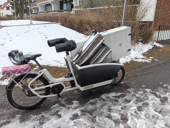 Urban Arrow Family bike loaded with a peice of white furniture (a shoe cabinet) and two steel bins. 

The bike has a pink cushioned seat on the back for a child to ride on. Behind the bike there is snow on the ground.