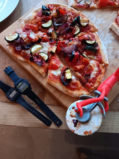 Vegetable pizza, two digital watches and a pizza cutter that looks like a monkey on a unicycle