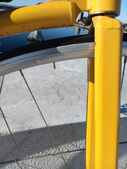 Close up of a penny farthing wheel showing a missing spoke. 

The sculpture, "She Lies" is viewable between the wheel and the yellow frame of the penny farthing. Giving away that this picture was taken from the top of the Oslo Opera house.