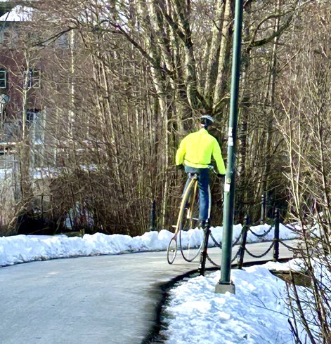 Picture of a person on a penny farthing bike, taken from behind. The person is wearing a fluorescent jacket. There is snow on the ground.