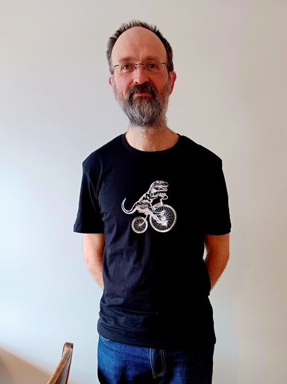 Picture of a man with s grey beard looking at the camera.  He is wearing a black t-shirt with a dinosaur riding a penny farthing. 