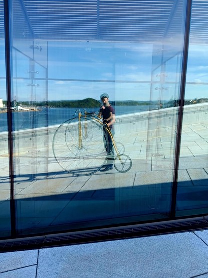 Reflection in multiple glass windows of a man taking a selfie (via the windows). He is standing on a steeply slanted, white surface and holding a yellow, "penny farthing" bicycle. In the bacground you can see the Olso fjord on a sunny day.