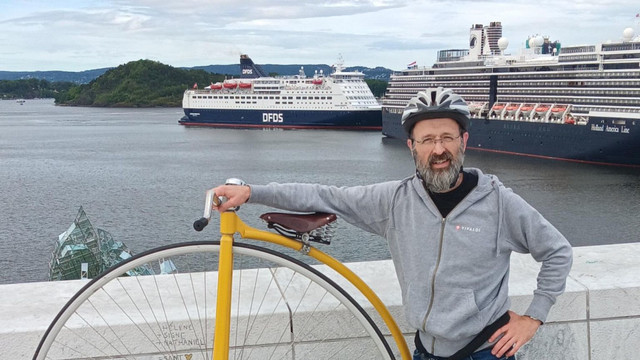 Man with a Vivaldi Browser hoody standing next to a yellow, large wheeled ("Penny Farthing") bicycle. Behind him is the Oslo fjord and two ferries.