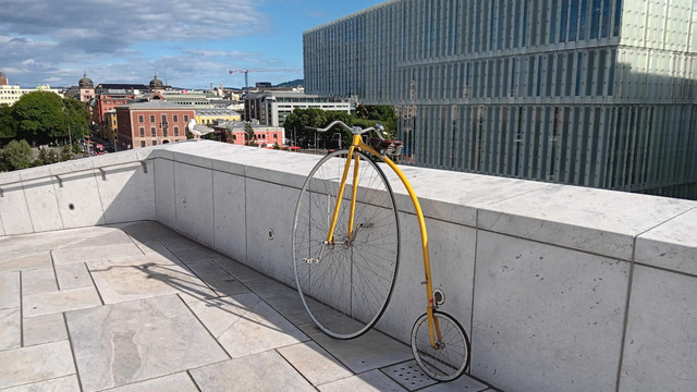 Yellow, penny farthing bicycle leaning against a white stone wall with a city scape in the background.