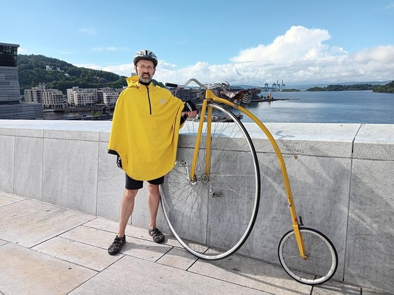 Man in a bright yellow rain cape, standing next to a large yellow "penny farthing" bicycle next to a white stone wall. The picture is taken at the top of the Oslo opera house, overlooking the Oslo fjord.