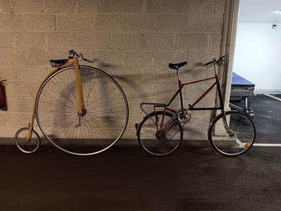 A yellow penny farthing bicycle and a black and red tallbike leaning against a wall in a garage. Their seat height appears to be the same. 