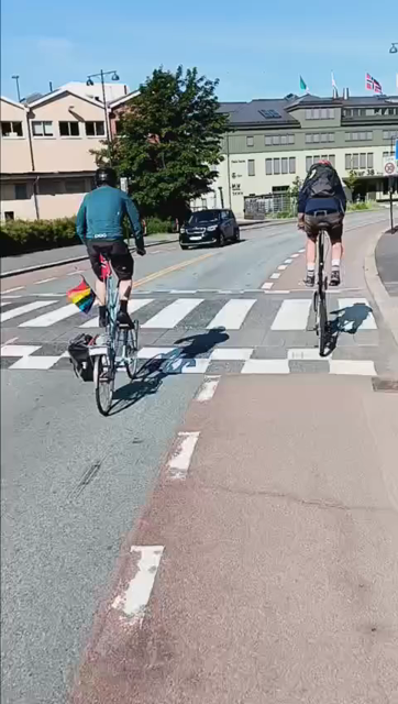 Two people on tallbikes cycling side by side. One of the bikes has both Norwegian and Pride flags.