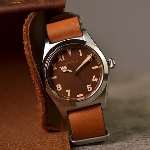 Baltany branded watch with a milk chocolate brown face featuring a 'Mercedes' style hour hand, while the minute hand is sword-shaped with its sides angling outwards. The hourly markings use the vintage Rolex 'error-proof' layout (a.k.a. the 'California dial'). As such the 12 is a downward facing triangle, 3/6/9 are horizontally aligned batons. For the remaining numbers, the upper ones are roman numerals, while the lower ones are arabic numerals. The watch has a light brown leather strap and a p…