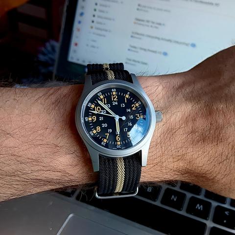 Analog watch in field watch style. Black dial with orange/yellow numerals for the main hours. There is also an inner ring for the 13-24 hours and an outer minute ring with 5 minute markers. Steel case, prominent crown. On a black NATO strap with a yellow stripe down the middle.