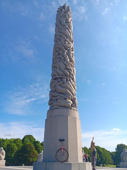 A red 29 inch wheeled unicycle in front of the Monolith scuplture (Monolitten). A 17 metre high carved stone column made up of naked human bodies of various ages and genders that is part of Frogner Park in Oslo