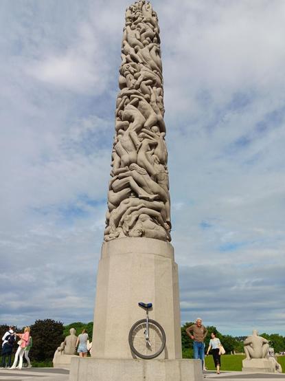 A silver 36 inch wheeled unicycle in front of the Monolith scuplture (Monolitten). A 17 metre high carved stone column made up of naked human bodies of various ages and genders that is part of Frogner Park in Oslo. Cloudy sky during day time.
