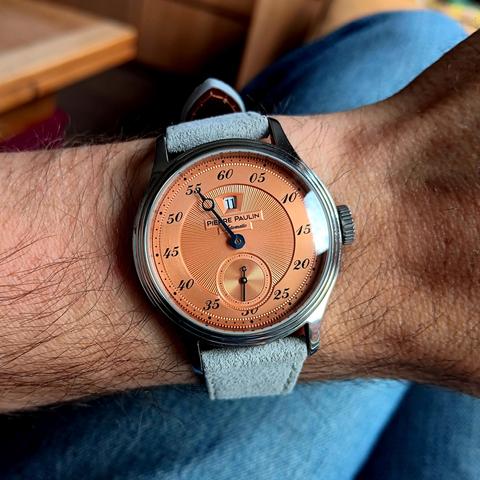 Watch with copper/salmon dial and hour jump complication, showing the time as 11:55. On a soft gray leather strap. 