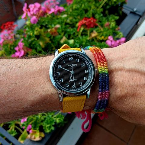 Widdershins running watch with a black dial and yellow leather strap. It shows the time as 5:10[PM]. On the same wrist is a rainbow band. In the background are some flowers. 