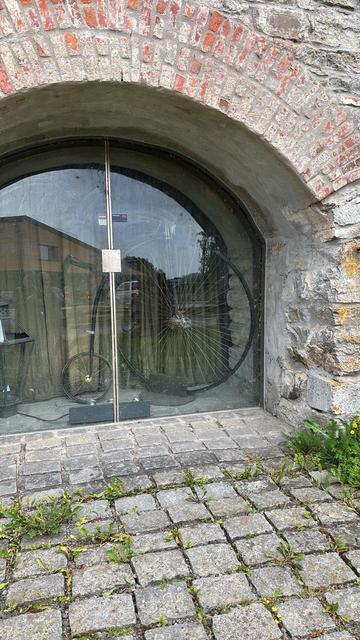 An old brick archway with a glass paned doors nestled into a cobblestone walkway. Framed inside is a penny farthing bicycle (a classic style of bike with a massive front wheel and a tiny rear one). It's positioned in front of a dark gray curtain, to hide whatever is behind it. To the left, there is a rustic looking lantern. Just in front is a set of lights, presumably to light it up at night. A small sign on the glass doors warns "ALARM".